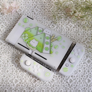 May include: White Nintendo Switch console with a custom skin featuring green and white cartoon cats and a green and white dessert design. The console is shown with two white Joy-Con controllers.