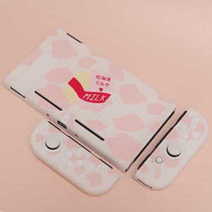 Puede incluir: Una piel de consola Nintendo Switch rosa y blanca con un diseño de estampado de vaca. La piel presenta un diseño de cartón de leche con el texto "北海道ミルク MILK" en japonés e inglés.