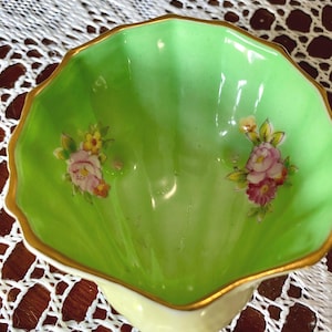 May include: A small, scalloped-edge bowl with a light green interior and a cream-colored exterior. The rim is trimmed with gold. Floral designs in pink, yellow, and green are painted on the interior. A decorative piece for serving or display.
