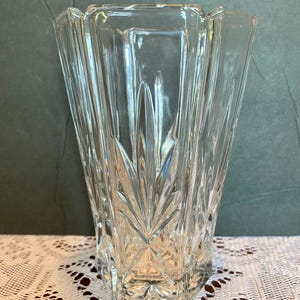 May include: A clear glass vase featuring a faceted, geometric design with vertical panels and a starburst pattern at the base. The vase has a wide, flared opening and a thick, sturdy bottom, showcasing a transparent, reflective surface throughout.