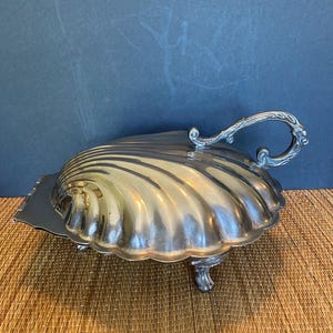May include: A silver-toned metal serving dish shaped like a large scallop shell. The dish features a hinged lid with a decorative curved handle and is supported by three small, ornate feet. The surface has a polished, reflective finish.