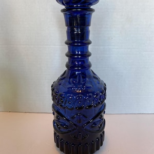 May include: A cobalt blue glass bottle with a decorative stopper. The bottle has a long neck and a textured, patterned body. The stopper has a swirl design. The bottle is likely vintage and is suitable for display.