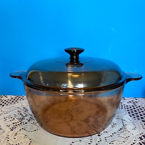 May include: A round, amber-colored glass casserole dish with a matching translucent lid. The dish features two integrated side handles and a central knob on the lid. The glass is smooth and transparent, showing a warm brown tint throughout the piece.