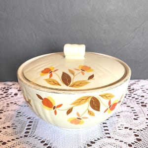 Op de afbeelding: Crème keramische kom met bijpassende deksel. Versierd met een bloemmotief in oranje, geel en bruin. De deksel heeft een kleine, rechthoekige handgreep. Geschikt voor het serveren van voedsel.