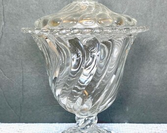 Puede incluir: Un cuenco de cristal transparente para caramelos con tapa. El cuenco tiene una base de pedestal y un diseño en espiral en el cuerpo. La tapa tiene un diseño de remolino y un adorno decorativo. El cuenco se utiliza para servir caramelos.