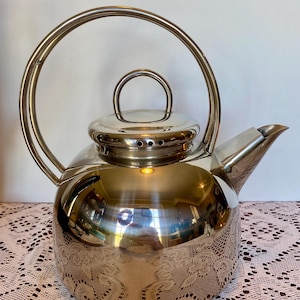 May include: A shiny, stainless steel teapot with a large, arched handle and a small, rounded lid handle. The teapot has a spout and a decorative, etched pattern around the base. The teapot is sitting on a white lace tablecloth.