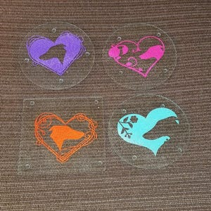 May include: Four glass coasters with a heart-shaped design. Each coaster features a different colored silhouette of a bird or animal inside the heart. The colors are purple, pink, orange, and blue.
