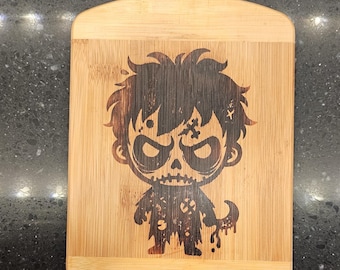 May include: A wooden cutting board with a burnt-in design of a cartoon zombie with a skull face. The zombie is wearing a tattered shirt and pants.