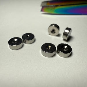Op de afbeelding: Vijf kleine, ronde, zilverkleurige metalen schijfjes met een gladde, reflecterende afwerking. Elke schijf heeft een gecentreerd, verzonken gat. Een deel van een regenboogkleurig, iriserend metalen gereedschap is rechtsboven zichtbaar.