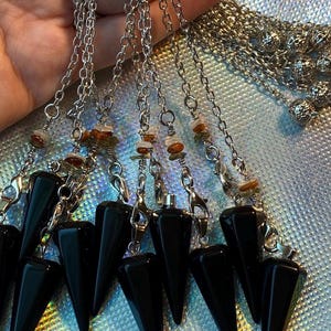 May include: A collection of black, cone-shaped pendulums with silver chains and clasps. Each pendulum is adorned with small, colorful gemstones. The pendulums are held against a shimmering, iridescent background.