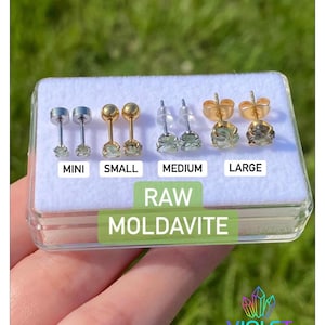 Puede incluir: Cuatro pares de pendientes de botón de moldavita en bruto dispuestos en una caja expositora de plástico transparente. Los pares están etiquetados como MINI, SMALL, MEDIUM y LARGE. El texto RAW MOLDAVITE aparece claramente. Logo VIOLET VESSEL.