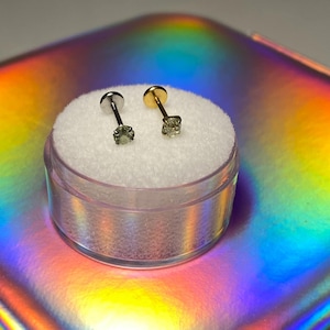 May include: Two small stud earrings with a gemstone, one silver and one gold, are displayed in a clear plastic container with a white, fluffy interior. The earrings sit on a reflective, iridescent surface.