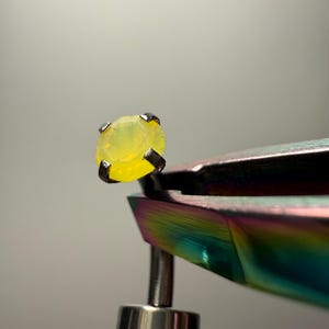 May include: A small, round, faceted yellow gemstone set in a four-prong silver-coloured metal stud earring base. The earring is held by iridescent, rainbow-coloured metal tweezers, showcasing the bright, translucent yellow stone and its setting.