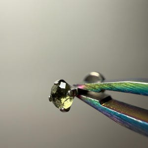 Puede incluir: Una pequeña piedra preciosa facetada de color verde oliva sostenida por unas pinzas metálicas iridiscentes. La piedra tiene un aspecto translúcido y texturizado con inclusiones internas visibles, asegurada en un engaste metálico simple de cuatro garras.
