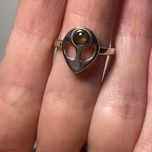 Puede incluir: Anillo de plata con diseño de cabeza de alienígena, con una pequeña gema redonda de color verde oliva. El anillo tiene una banda sencilla. Se ven anillos adicionales con piedras transparentes en otro dedo.