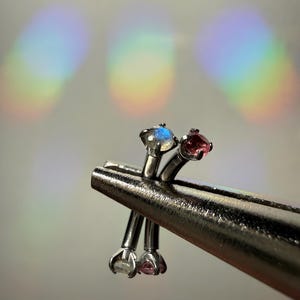Puede incluir: Primer plano de cuatro piercings de nariz plateados, cada uno con una pequeña gema de colores. Las gemas son en tonos rosa, transparente y azul. Los piercings están sujetos por una herramienta plateada sobre un fondo borroso.