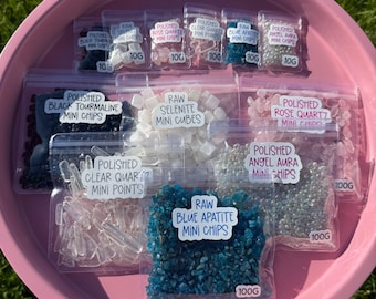 Puede incluir: Una bandeja rosa llena de pequeñas bolsas de plástico transparente que contienen varias piedras preciosas pulidas y en bruto. Las bolsas están etiquetadas con el tipo de piedra, como "Black Tourmaline Mini Chips". La bandeja está sobre hierba. Medidas en gramos.