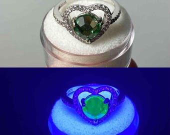 May include: A silver ring with a heart-shaped design. The ring features a large, round, green gemstone at the center, surrounded by a heart-shaped frame of smaller, clear stones. The band is also adorned with small stones.