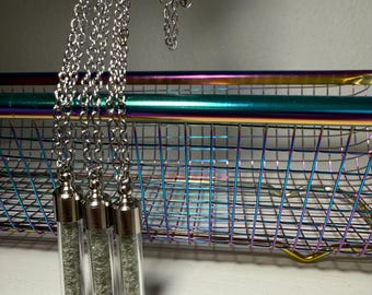 Op de afbeelding: Drie zilverkleurige kettingen met hangers van doorzichtige glazen flesjes gevuld met kleine, textuurrijke, metaalkleurige deeltjes. Elke hanger is afgewerkt met een gepolijste zilverkleurige metalen dop en hangt aan een bijpassende schakelketting.