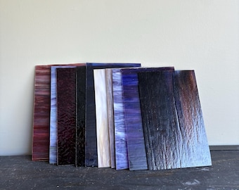 May include: A collection of rectangular stained glass sheets in various colors, including shades of red, purple, black, white, and blue. The glass has a textured appearance and is arranged vertically against a neutral background.
