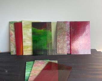 May include: A collection of rectangular stained glass sheets in various colors including red, green, yellow, and pink. Some sheets feature solid translucent tones, while others display marbled or textured patterns with swirling color variations.