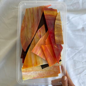 May include: A clear plastic rectangular container filled with assorted irregular shards of stained glass in shades of orange, red, yellow, and brown. The glass pieces feature marbled and streaked patterns with varying levels of translucency.