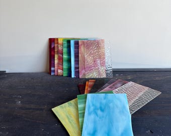 May include: A collection of colorful, square glass tiles. The tiles display a variety of colors, including red, yellow, blue, green, and pink. Some tiles have textured surfaces. The tiles are arranged on a dark surface.