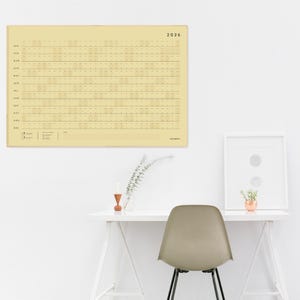 May include: A large, rectangular 2026 calendar in a pale yellow hue hangs on a white wall. The calendar features a grid layout with horizontal lines for each day. A white desk with a chair and framed art are below.