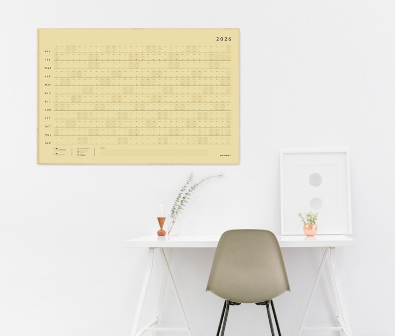 May include: A large, rectangular 2026 calendar in a pale yellow hue hangs on a white wall. The calendar features a grid layout with horizontal lines for each day. A white desk with a chair and framed art are below.