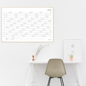 May include: A framed 2026 year calendar with a minimalist design. The calendar is white with gray grid lines and text, and is hanging on a white wall. A desk with a chair and decorative items are in front of the wall.