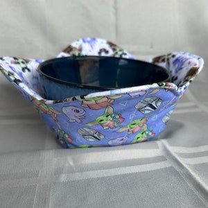 May include: A blue bowl with a white rim sits inside a fabric bowl cozy. The cozy is made of a light blue fabric with a white background and features a repeating pattern of cartoon characters from the Star Wars franchise.