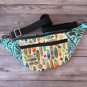 May include: A blue and brown fanny pack with a white background featuring colorful surfboards and the text "Ride the Waves". The fanny pack has two zippered compartments and a black adjustable strap.