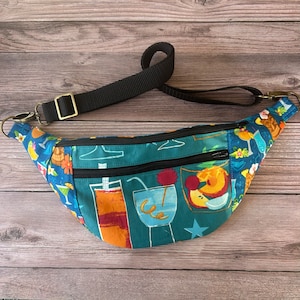 May include: A teal blue fanny pack with a black adjustable strap. The fanny pack has a tropical print featuring colorful cocktails and a star.
