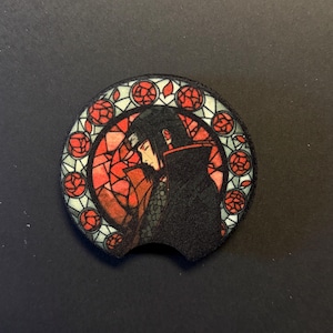 Puede incluir: Posavasos negro para coche con diseño de vidriera. Presenta un personaje con capa negra y detalles rojos. El posavasos es circular con un borde curvado, diseñado para encajar fácilmente en un portavasos de coche.