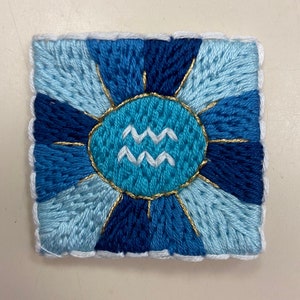 May include: A blue and white embroidered square with a gold sunburst design and the astrological symbol for Aquarius in the center.