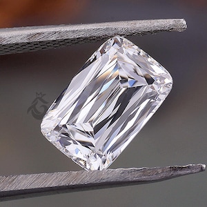 May include: A clear, colourless, rectangular emerald-cut gemstone held by metal tweezers. The stone features sharp, geometric facets that reflect light, creating a brilliant, multi-faceted appearance with a small logo mark visible on the surface.