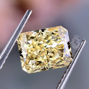 May include: A radiant-cut yellow gemstone held by silver-toned metal tweezers. The stone features a multifaceted surface that reflects light, creating a brilliant, sparkling appearance with a distinct rectangular shape and a small logo mark.