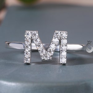 May include: A silver ring featuring the letter "M" encrusted with small, sparkling diamonds. The ring is set against a muted blue-gray surface, with a blurred green plant in the background. The ring's design is simple and elegant.