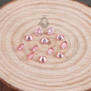 Peut inclure: Une collection de petites pierres précieuses facettées rose clair disposées en cercle sur une tranche de bois ronde et claire, avec des cernes visibles et une écorce sombre et texturée. Un petit logo est imprimé au-dessus des pierres.