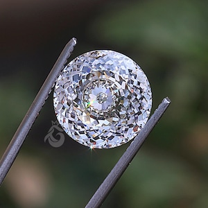 May include: A round, colorless, faceted gemstone with a brilliant cut, displaying multiple triangular and kite-shaped facets that reflect light. The stone is held by metal tweezers and features a small logo mark on its lower left side.