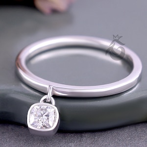 May include: A silver ring with a square, clear gemstone pendant. The ring band is thin and polished, and the gemstone is set in a square bezel. The ring is displayed on a dark gray surface, with blurred pink flowers in the background.