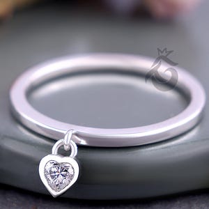 May include: A silver ring with a heart-shaped pendant. The pendant is a clear, sparkling gemstone set within a heart-shaped frame. The ring band is thin and polished, and the pendant hangs freely from the band.