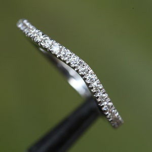 May include: A curved silver-toned ring band featuring a row of small, clear, round-cut stones set along the top surface. The metal has a polished, reflective finish, and the band follows a gentle wave-like contour throughout its entire length.
