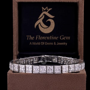 Op de afbeelding: Een zilverkleurige tennisarmband met een doorlopende rij vierkant geslepen, heldere stenen. De armband wordt gepresenteerd in een bruin fluwelen doosje met de tekst The Florentine Gem A World Of Gems & Jewelry in goud op de binnenzijde.