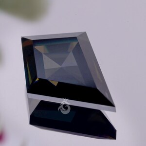 May include: A kite-shaped gemstone with a dark, deep blue and black hue, featuring a faceted surface that creates geometric light reflections. A small white logo depicting a stylised crown or leaf shape is visible on the lower facet of the stone.