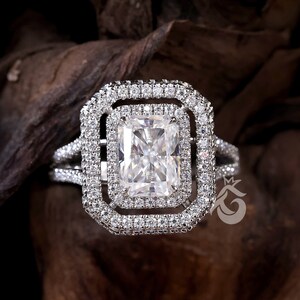 Puede incluir: Un anillo de diamantes con un diamante central de talla radiante, rodeado por dos halos de diamantes más pequeños. La montura también está adornada con diamantes pavé, creando un efecto deslumbrante. El diseño es elegante y llamativo.
