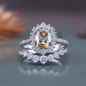 May include: A silver ring set featuring a central cushion-cut champagne-coloured gemstone surrounded by a halo of small round white stones. The band is accented with additional small white stones, and a matching curved band sits below the main ring.