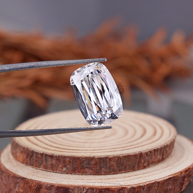 May include: A clear, colorless, cushion-cut gemstone with a brilliant faceted surface. The stone is held by metal tweezers, displaying sharp internal light reflections and a rectangular shape with rounded corners, showcasing high clarity and transparency.