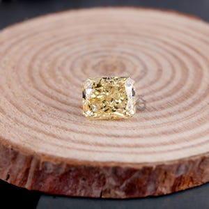May include: A radiant-cut yellow gemstone with a faceted surface that reflects light, displaying a bright golden hue. The stone features a rectangular shape with clipped corners and is positioned centrally on a circular, light-colored wood slice.