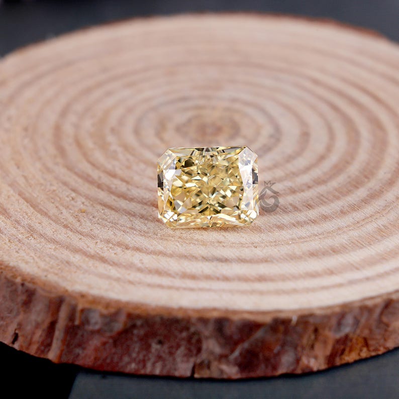 May include: A radiant-cut yellow gemstone with a faceted surface that reflects light, displaying a bright golden hue. The stone features a rectangular shape with clipped corners and is positioned centrally on a circular, light-colored wood slice.